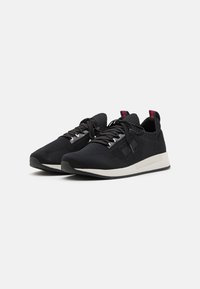 Paar schwarze gestrickte Low-Top-Sneaker mit weißen Sohlen, schwarzen Schnürsenkeln und roten Zuglaschen auf einem weißen Hintergrund.