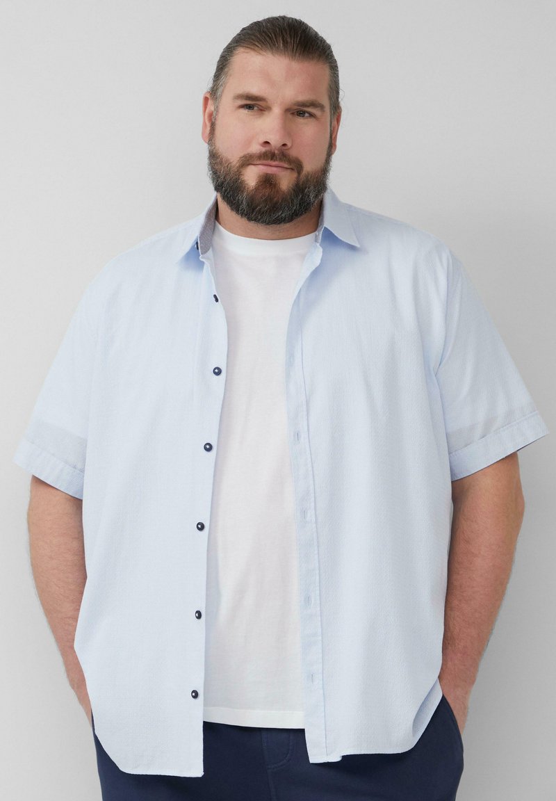 Korte mouwen lichtblauw overhemd met een gemêleerd patroon, voorzien van donkere knopen en een wit T-shirt eronder.