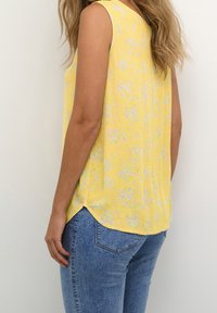 Mouwloze gele blouse met een bloemmotief, met een ronde zoom en zijsplitten, gecombineerd met blauwe spijkerbroek.