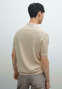 Beige strikket polo t-shirt med korte ærmer, prydet med et tekstureret mønster og ribbet krave. Bagside viser en tætsiddende design og minimalistiske syninger.