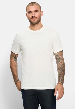 Uomo con capelli corti e braccia tatuate che indossa una maglietta bianca semplice a maniche corte e pantaloni scuri, in piedi davanti a uno sfondo chiaro.
