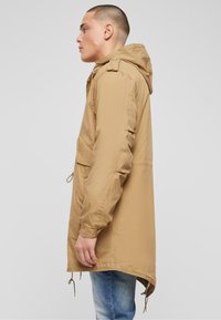 Giacca cappuccio marrone chiaro con vestibilità comoda, caratterizzata da lacci al fondo, due tasche frontali e un tessuto leggero e liscio.