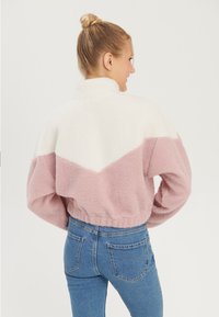 Veste en polaire avec un col blanc et un corps rose, présentant un design en chevrons. Style cropped avec ourlet élastique, portée avec un jean bleu.
