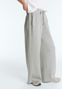 Person iført lysegrå sweatpants med vide ben og snøre i taljen, hvide sneakers og en hvid skjorte lagt ned i bukserne.