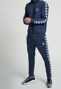 Navy tracksuit made of soft fabric, featuring a zip-up jacket with white chevron stripes on sleeves and matching tapered pants. Black athletic shoes.