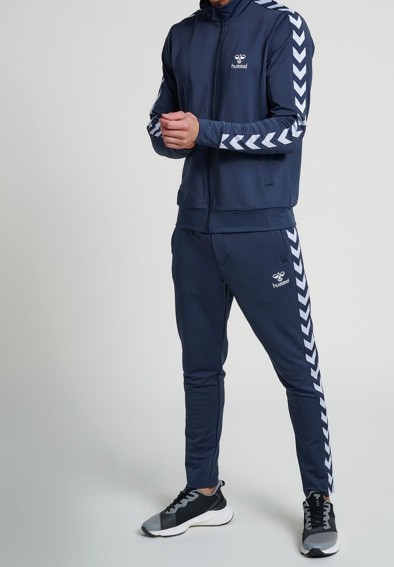 Navy tracksuit made of soft fabric, featuring a zip-up jacket with white chevron stripes on sleeves and matching tapered pants. Black athletic shoes.