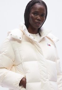 Μπουφάν puffer σε ανοιχτό κρεμ χρώμα, με κουκούλα, ραμμένο καπιτονέ μοτίβο και κλείσιμο με κουμπιά πατιλέτα. Μπαλώμα με λογότυπο στο στήθος.