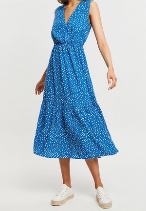 Kobieta w niebieskiej sukience midi bez rękawów w białe grochy i białych platformowych trampkach, stojąca z lekko wyciągniętymi ramionami.