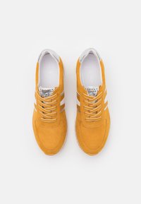 Zapatillas de ante amarillas con rayas blancas, detalles texturizados, ojales metálicos y cordones redondos. Interior suave y suela acolchada.