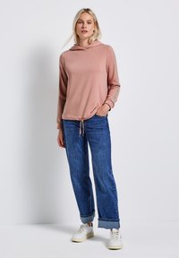 Sudadera rosa clara con cordones, mangas largas y rayas laterales, combinada con vaqueros de pierna ancha de color azul medio y zapatillas blancas.