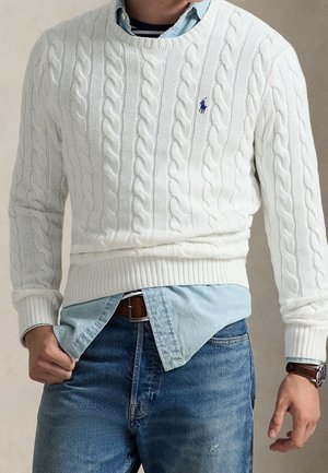 Mand iført hvid kabelstrikket sweater over lyseblå skjorte stoppet ned i blå jeans med brunt bælte og armbåndsur på venstre håndled.