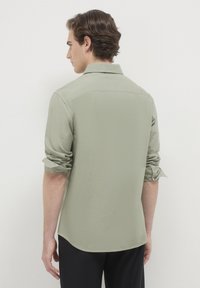 Uomo con capelli castani che indossa una camicia a maniche lunghe verde chiaro con le maniche arrotolate, girato di spalle contro uno sfondo chiaro e uniforme.