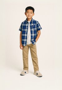 Cămașă cu mânecă scurtă în carouri albastre peste un tricou alb, pantaloni kaki și adidași deschiși cu accente bleumarin. Nasturi și buzunar pe piept vizibile.