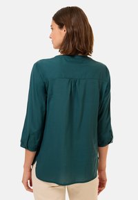 Blusa teal con maniche 3/4 e orlo arrotondato. Tessuto morbido e leggero con dettaglio plissettato sul retro e polsini delle maniche arrotolati.