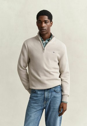 Jonge man met een beige quarter-zip trui over een blauw overhemd met kraag en blauwe jeans, staand tegen een effen lichte achtergrond.