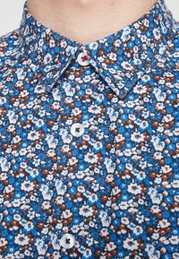 Gros plan sur le col et les boutons d'une chemise à motif floral bleu, présentant de petites fleurs blanches et brunes avec des boutons blancs.