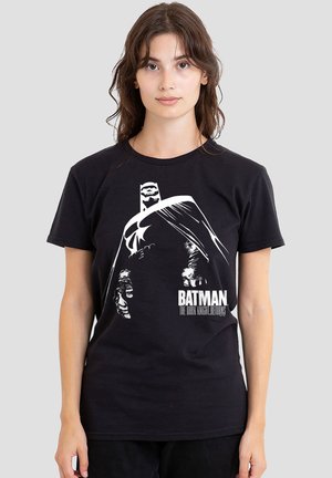 Young woman wearing a black T-shirt with a white graphic of Batman and text "BATMAN THE DARK KNIGHT RETURNS" on a plain gray background.