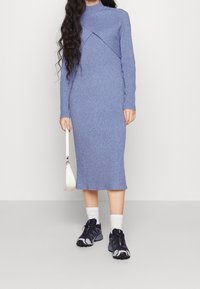 Vestido de punto acanalado azul claro con un cuello alto y diseño cruzado, combinado con zapatillas oscuras y un pequeño bolso blanco.