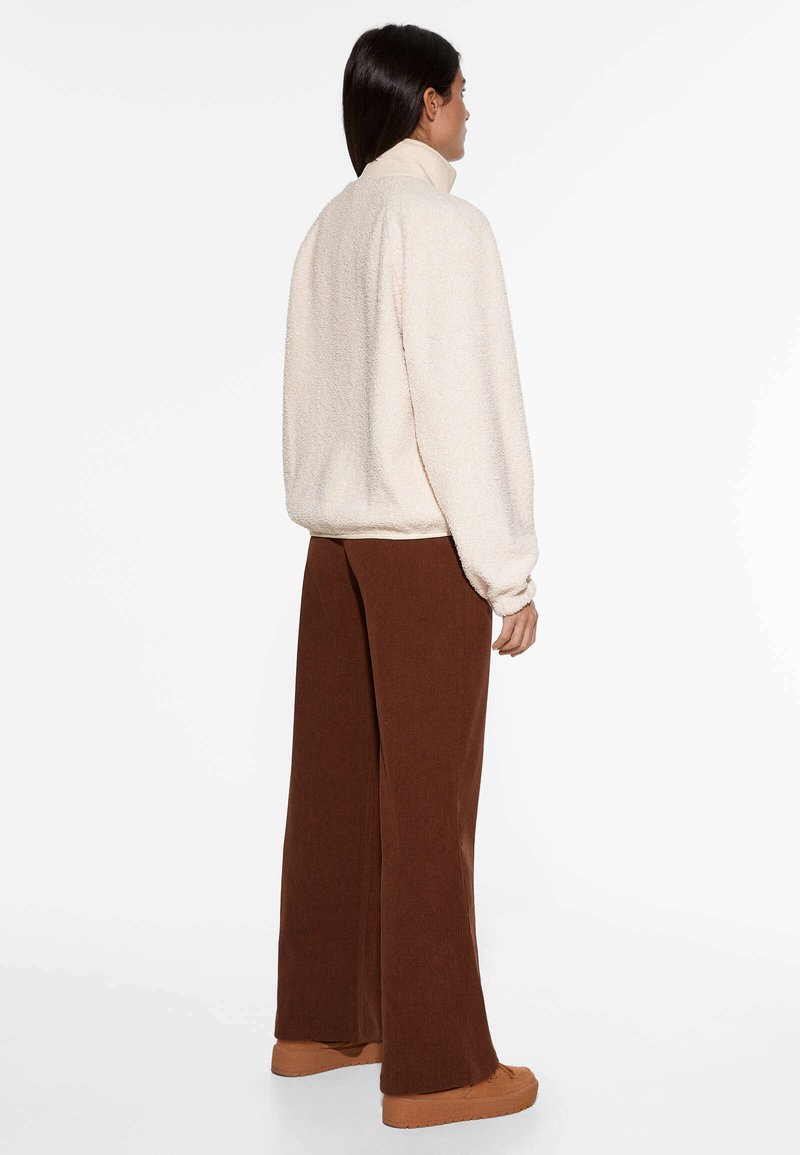 Crème-kleurig fleece sweater met een hoge kraag, gecombineerd met bruine wijde broek. De sweater heeft een gestructureerd, fluffy oppervlak.