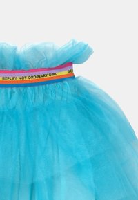 Jasnoniebieska spódnica z tiulu z falbanką w talii, będąca ozdobiona wielokolorowym paskiem z napisem "REPLAY NOT ORDINARY GIRL". Miękka tekstura.