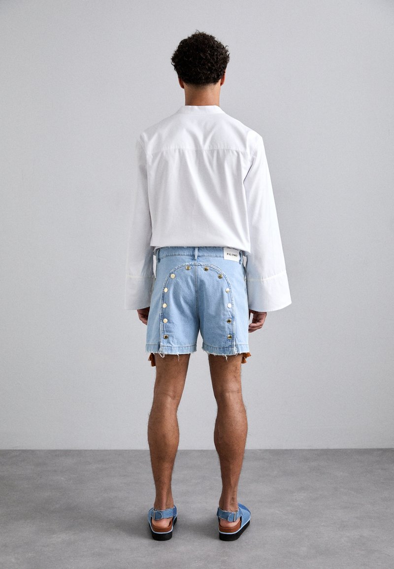 Shorts in denim azzurro chiaro con orli sfrangiati, dettagli con bottoni lungo i lati, e una camicia bianca a maniche lunghe, vista da dietro.