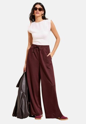 Frau mit ärmellosem weißen Oberteil, hoch taillierter, weit geschnittener maroonfarbener Nadelstreifenhose, Sonnenbrille, trägt schwarze Lederjacke und maroonfarbene Schuhe.