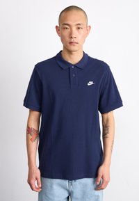 Námornícky modrá polokošeľa s golierom, dvoma gombíkmi a malým bielym logom Nike na ľavej strane hrude. Mäkký materiál, krátke rukávy.