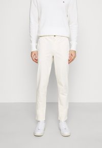 Osoba v bílých slim-fit kalhotách, bílém svetru s dlouhým rukávem a logem na hrudi, a bílých teniskách, stojí na bílém pozadí.