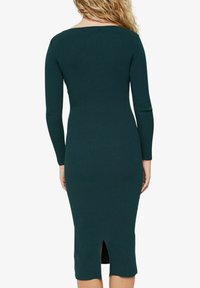 Robe côtelée vert foncé à manches longues, encolure bateau et fente haute à l'arrière, ajustée à la silhouette.