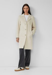 Manteau en laine beige avec un col à revers, un devant à bustier unique et deux poches latérales, associé à un pantalon gris évasé et des baskets multicolores.