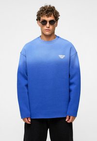 Blauwe sweatshirt met een ombre-effect, geribbelde ronde hals, verlaagde schouders en een klein wit logo op de borst, gecombineerd met ronde zonnebrillen.