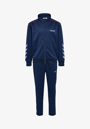 Ensemble de survêtement navy avec veste zippée et pantalon assorti, présentant des accents rouges et des motifs en chevron blancs sur les manches. Fabriqué en matériau lisse et léger.