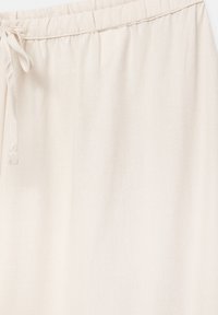 Pantalon en tissu beige clair avec une ceinture élastique et un cordon noué sur le côté gauche, représenté en gros plan.