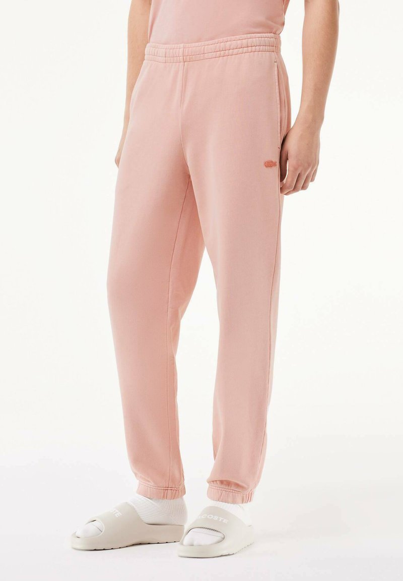 Pantalons de survêtement rose clair avec taille élastique et poignets côtelés. Le côté gauche présente un petit logo Lacoste brodé. Portés avec des claquettes blanches.