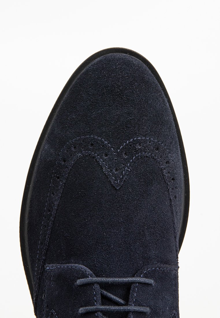 Chaussure brogue en nubuck bleu marine présentant un bout arrondi, des perforations décoratives et un laçage ; texture lisse avec des accents de couture contrastants.