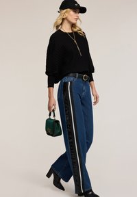 Czarny sweter z prążkowym wzorem, spodnie dżinsowe z wysokim stanem z cekinami i akcentami w paski, zielona torebka i czarne botki na obcasie. Casualowy strój.