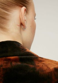 Pendiente plateado en forma de corazón llevado en una oreja perforada, con un pequeño tatuaje rojo que dice "oui" detrás de la oreja. El collar tiene un patrón oscuro y texturizado.