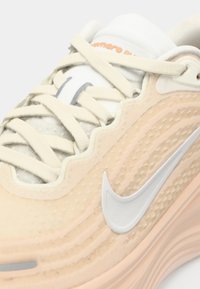 Sneaker bej cu partea superioară texturată, șireturi albe și logo-ul Nike argintiu. Prezintă un design ondulat și un guler căptușit pentru confort.