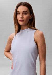 Νεαρή γυναίκα με ίσια καστανά μαλλιά, που φορά αμάνικο ανοιχτό λεβάντα τοπ, στέκεται μπροστά σε απλό γκρι φόντο.