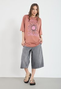 Overdimensioneret terracotta t-shirt med solgrafik, grå brede denimshorts og lyse sandaler med sorte såler og spænder.