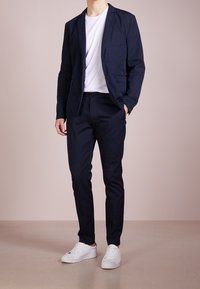 Granatowa marynarka z dwiema kieszeniami z przodu, zestawiona z pasującymi spodniami. Nosi się ją na białej koszulce i białych trampkach.