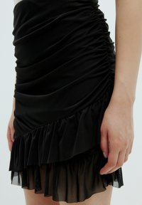 Zwarte jurk met gerimpelde textuur en gelaagde ruffles aan de zoom. De stof lijkt licht glanzend en heeft een soepele, lichte afwerking.