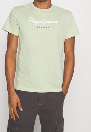 Mężczyzna ubrany w jasnooliwkową koszulkę Pepe Jeans London oraz ciemne spodnie cargo, z rękami w kieszeniach, pokazany od szyi do górnej części ud.