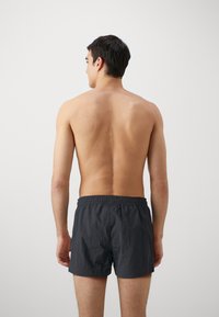 Costum de baie pentru bărbați, de culoare neagră, cu talie elastică, fabricat dintr-un material ușor, având o textură simplă. Văzut din spate.