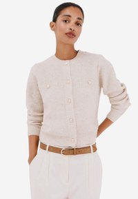 Cardigan in maglina beige con scollo rotondo, polsini a coste e due tasche frontali. Presenta grandi bottoni simili a perle e dettagli testurizzati.