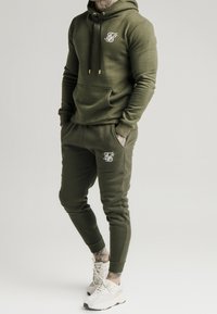 Olivgrüner Reißverschluss-Hoodie mit Fronttasche und weißem Logo, kombiniert mit passenden Jogginghosen. Beide haben gerippte Bündchen und eine weiche Textur.