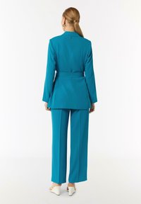 Turkusowa marynarka z paskiem w talii oraz szerokimi spodniami, wykonana z gładkiego materiału. Widok z tyłu ukazuje dopasowane szwy oraz minimalistyczny design.