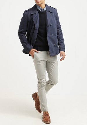 Uomo che indossa un caban blu navy, maglione nero, pantaloni grigio chiaro e scarpe eleganti in pelle marrone, in piedi con una mano in tasca.