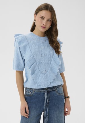 Nő világoskék, rövid ujjú kötött pulóverben fodrokkal és fonott mintával, kék farmerral és gyöngy fülbevalóval.