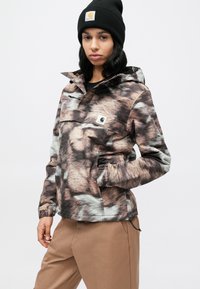 Pulloverjack met een bontachtige print in tinten bruin en grijs. Voorzien van een voorzak, verstelbare manchetten en een capuchon. Draagt beige broek.